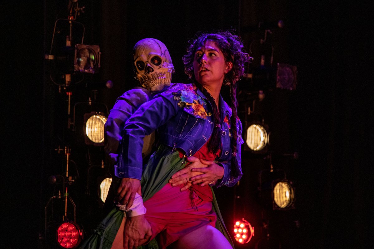 A woman with short brown curly hair, wearing a pink running vest and shorts, is dancing on stage with a skeleton in a suit. The skeleton has a skull face. The woman is standing in front of the skeleton and the skeleton has his hand on her waist and her leg.