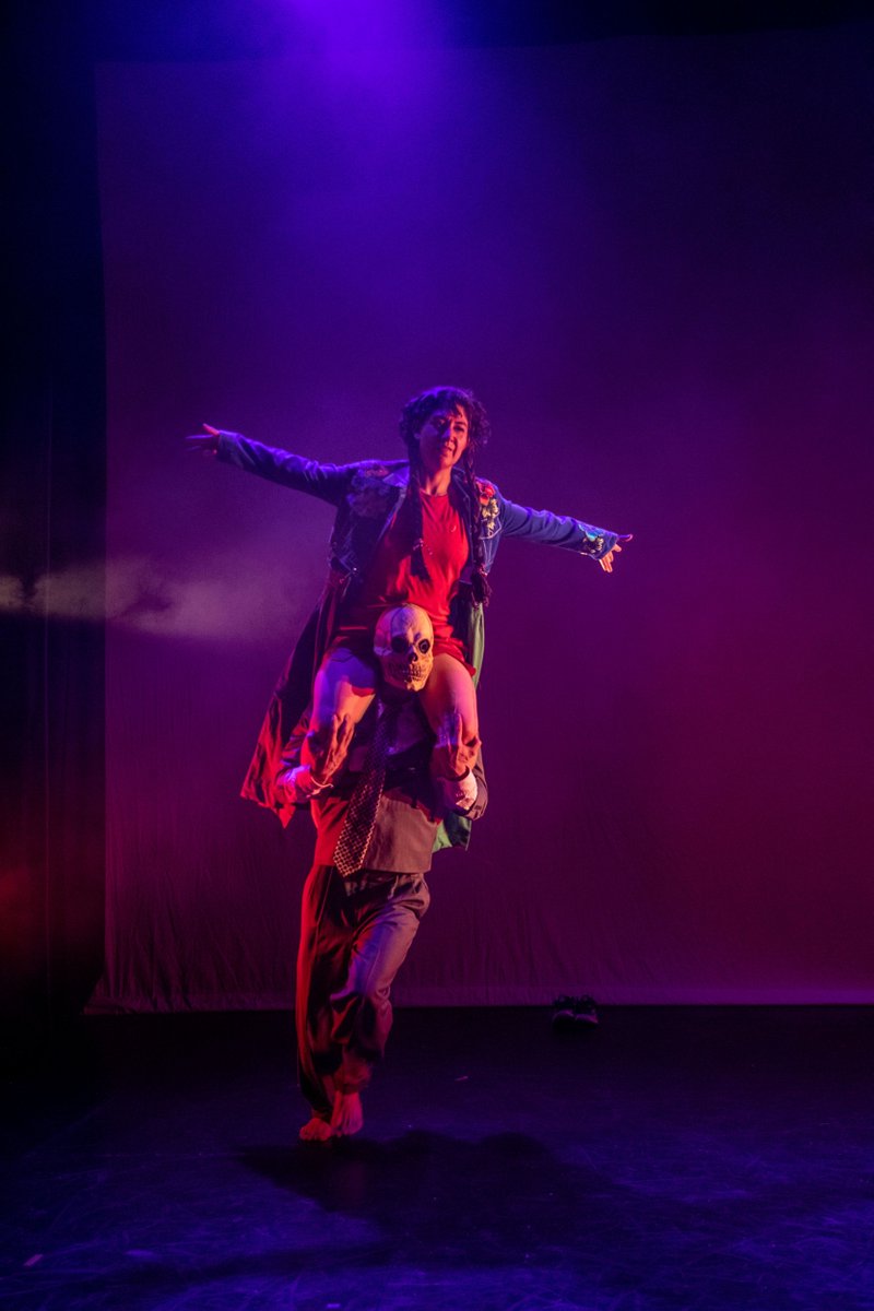 A woman with short brown curly hair, wearing a pink running vest and shorts, is dancing on stage with a skeleton in a suit. The skeleton has a skill face. The woman is sat on the skeletons shoulder.
