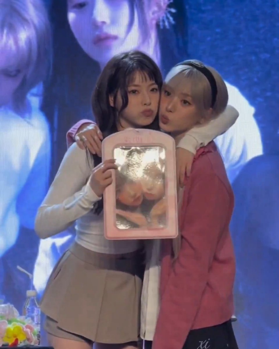 Two young women pose together making kissing faces while holding up a pink framed photo of themselves, in front of a large screen displaying an image.
