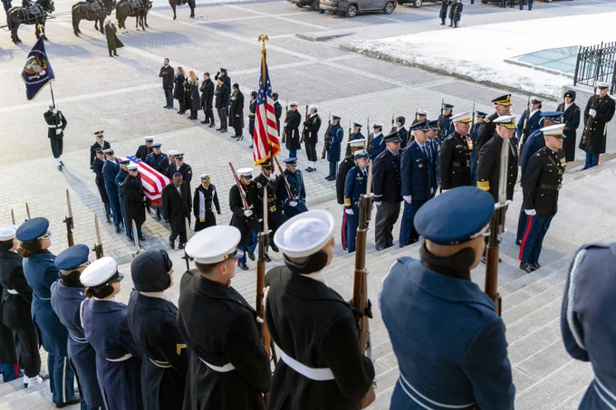 The Joint Chiefs escort uniformed pallbearers carrying the casket of the late President Carter up a large stair case.  