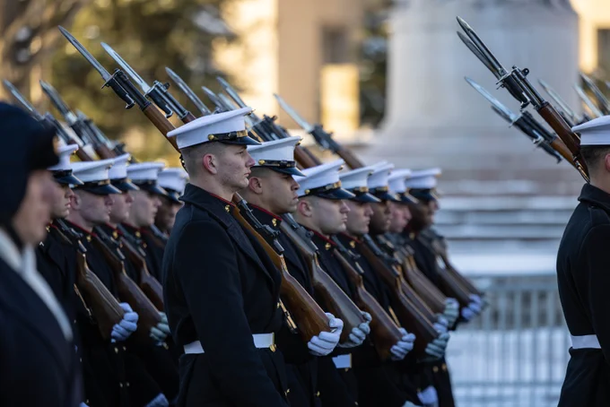 Uniformed servicemembers march in rows with muskets over their shoulders. 