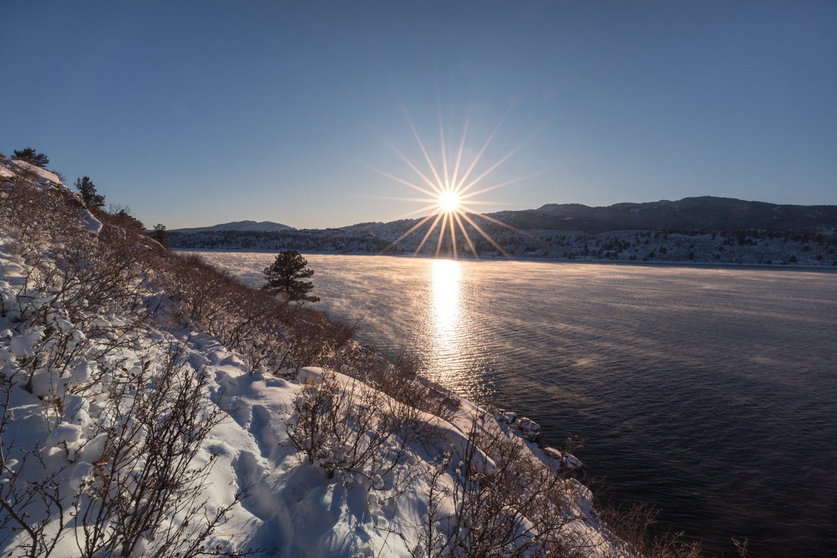 Horsetooth Reservoir blows in the wind surrounded by snowy foothills and shrubs, with a setting sun on the tip of the mountain ridge alongside Horsetooth Rock.