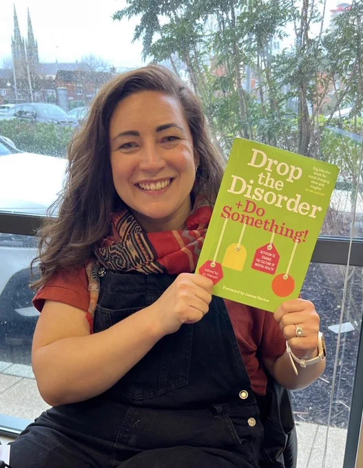 A woman holding a book with the title ‘Drop the Disorder + Do Something’.