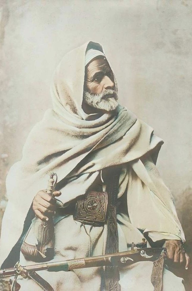 A Libyan Bedouin sat with a rifle on his lap and a powder horn tucked into his belt. Taken in Amsterdam in 1905.