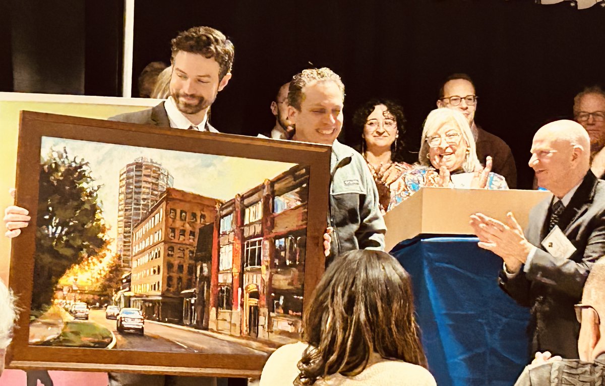 A group of people at an indoor ceremony or event pose with a large framed painting of a city street. One person holds a presentation box while another applauds. There is a small amount of text on clothing or a badge.