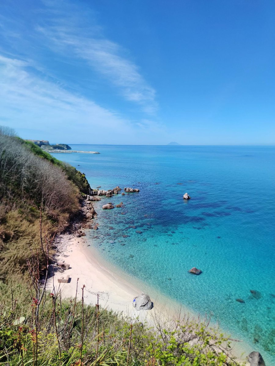 Una foto mozzafiato della spiaggetta di Michelino a Parghelia: la sabbia bianca e i suggestivi scogli creano un contrasto con il mare cristallino che sfuma dal turchese al blu profondo 🏖️🌊. Sullo sfondo, il porto di Tropea si staglia elegante ai piedi della città arroccata 🛥️,