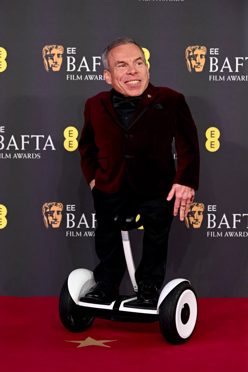 Warwick Davis poses for photos on the BAFTA Film Awards 2025 red carpet. He is wearing a burgundy suit blazer, and black pants and is standing on a motorised wheel device. He is looking away from the camera and smiling, with one hand in his pocket. 