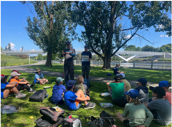A group of youth sitting on grass beside the Rideau Canal listening to two presenters