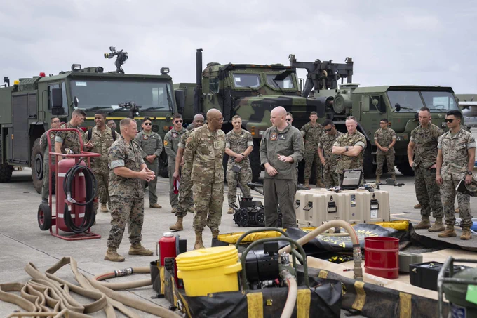 Chairman of the Joint Chiefs General CQ Brown, Jr. standing with a group of uniformed marines in front of three military-owned vehicles; one servicemember is speaking and gesturing to machine parts and other equipment sitting in the foreground.