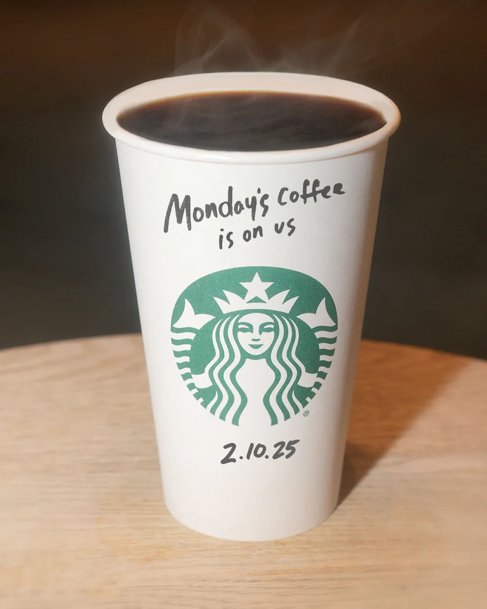 A tall Starbucks coffee sits on a table with "Monday's coffee is on us" and "2.10.25" written in Sharpie on it.