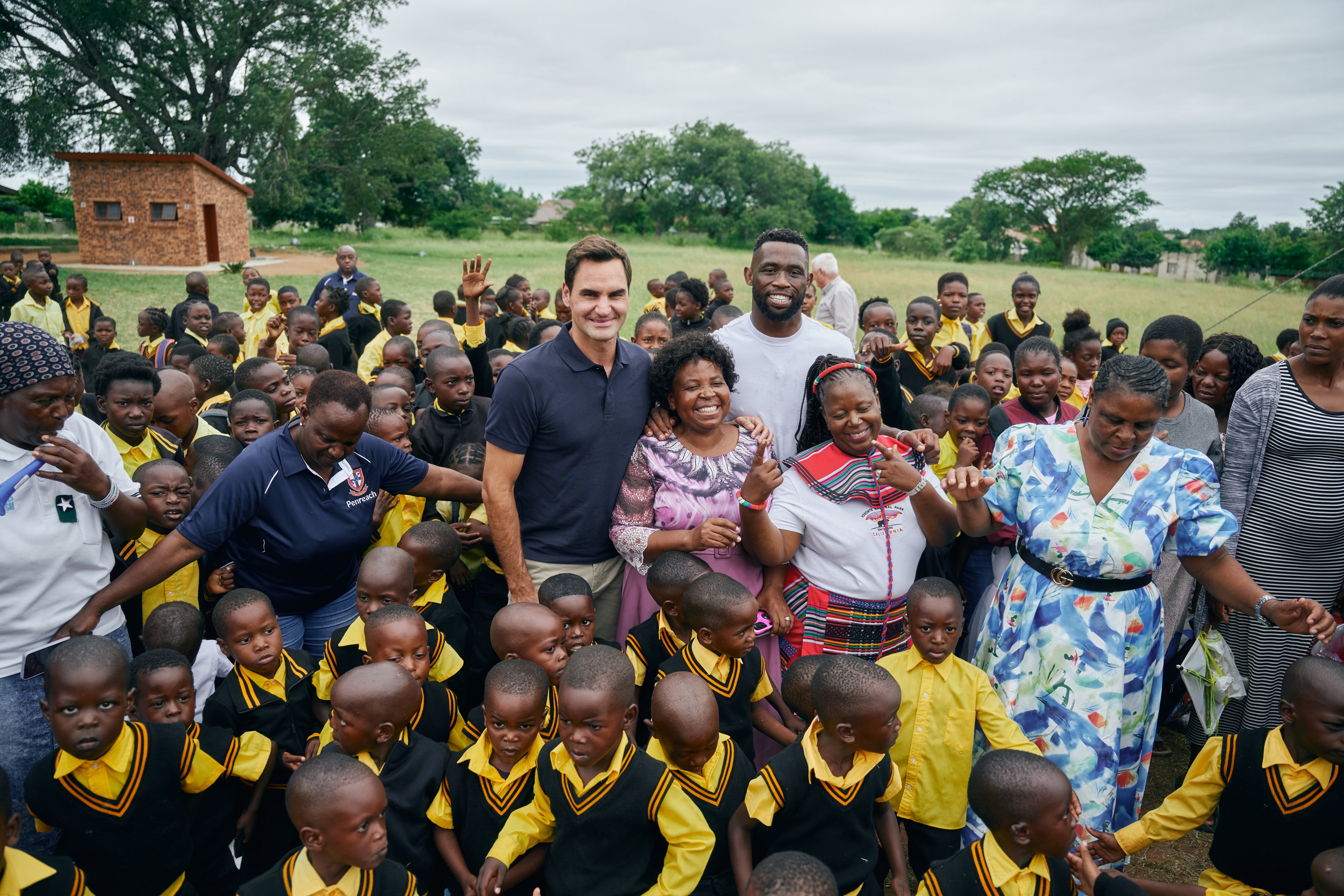 Roger Federer and Siya Kolisi surrounded by uniformed preschool children and staff. A green field and trees can be seen in the background.