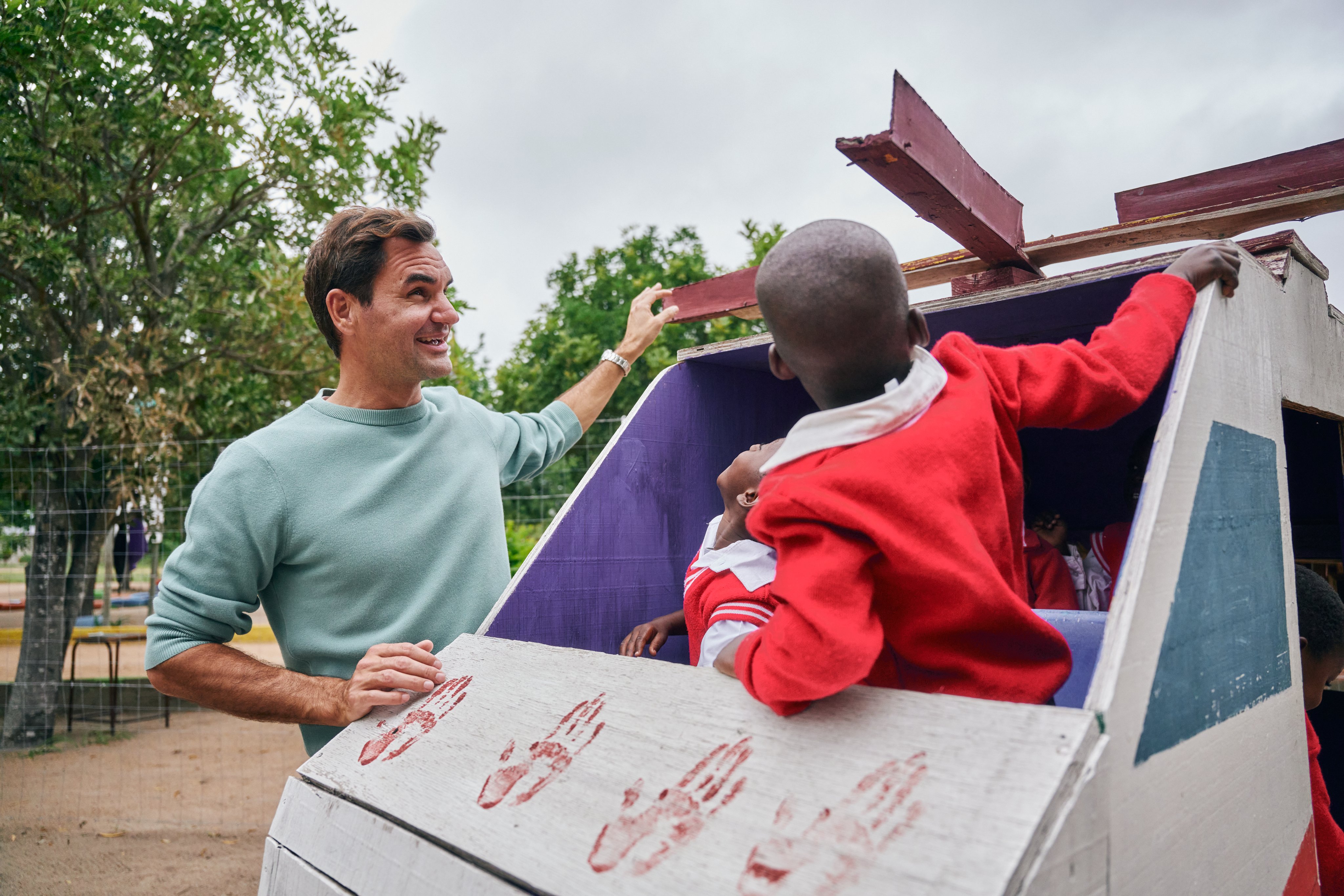 Roger Federer examines a playground element in the shape of a helicopter while two preschool children in the cockpit look on.