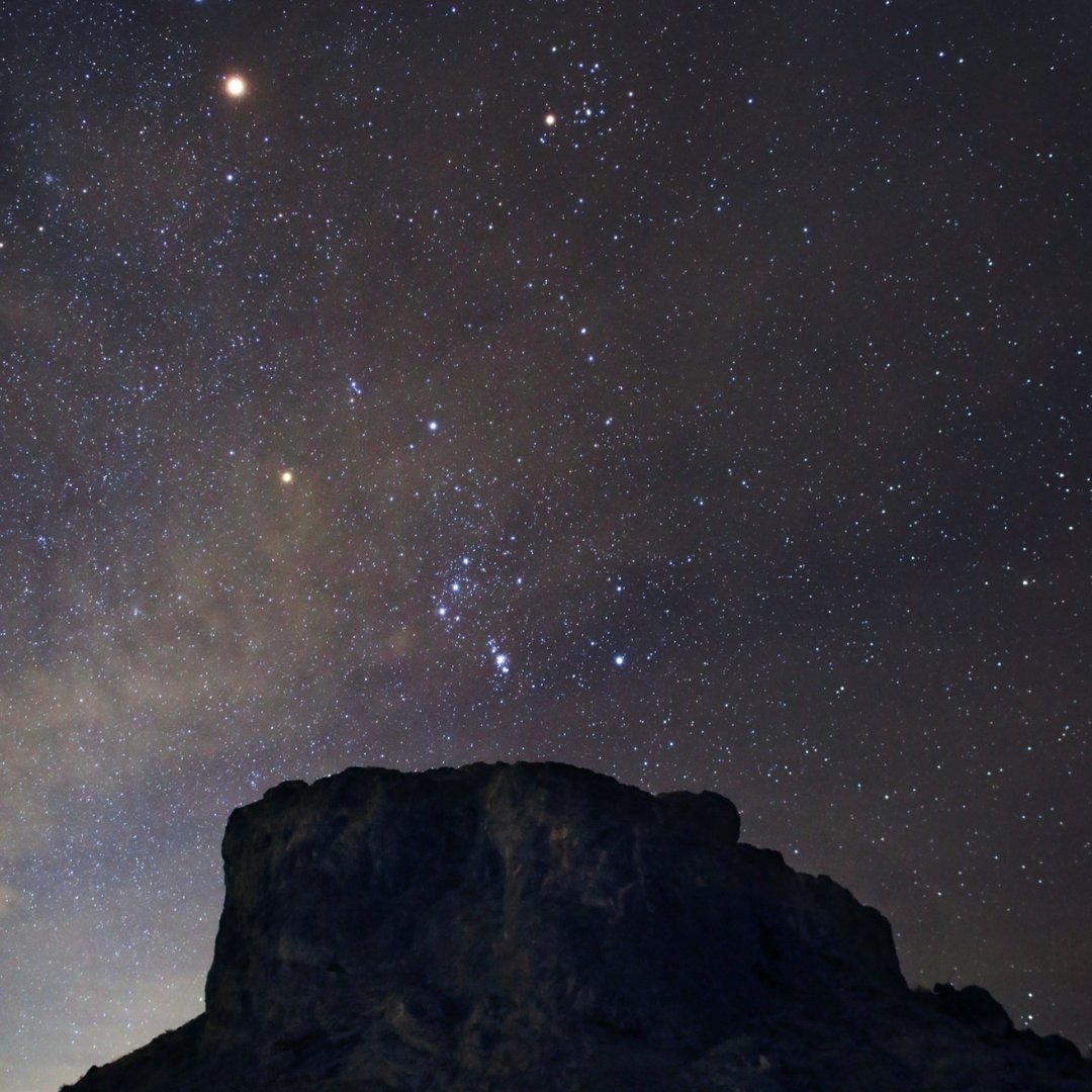 Estrellas y planetas brillantes y coloridos aparecen en un cielo nocturno nebuloso sobre la silueta oscura de un acantilado rocoso de Utah. 