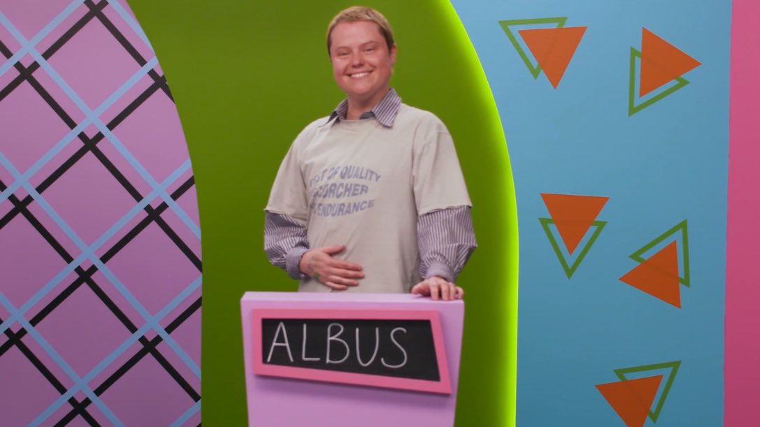 Ally Beardsley on Make Some Noise on Dropout, wearing a white graphic shirt over a lavender stripe button up, standing at a pink game podium that reads “Albus” in chalk, in front of a multi colorful retro stage backdrop.