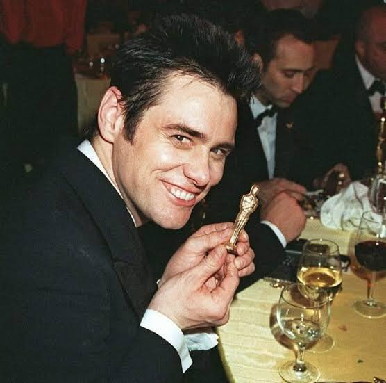 Hermosa foto de Jim Carrey con un pequeño Oscarcito mientras de fondo se ve a John Travolta.