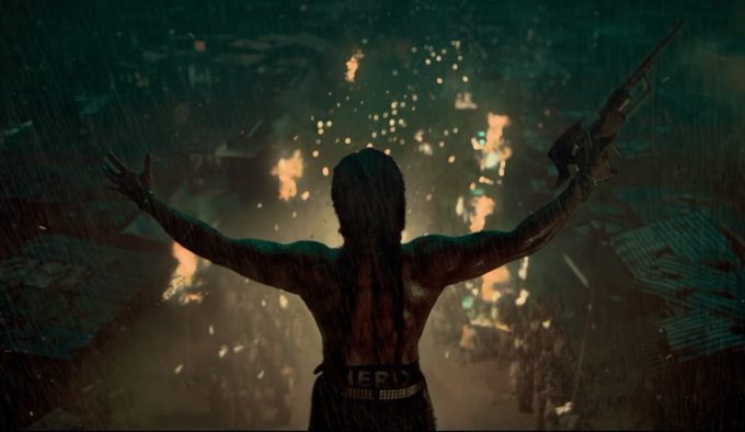 The image depicts a dramatic scene from the film "The Paradise," which is described as India's version of MadMax. The central figure, likely Nani, is shown from behind with arms raised, holding a weapon in one hand, symbolizing leadership and defiance. The setting is chaotic, with fires burning in the background, suggesting a battle or uprising. The context provided indicates that this film is set in Secunderabad during the 1980s, focusing on a marginalized tribal group fighting for their rights. The composition emphasizes the protagonist's silhouette against the fiery backdrop, highlighting the intensity and significance of the struggle depicted.