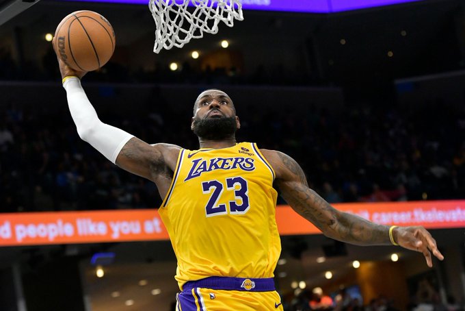 The image shows LeBron James, a prominent basketball player for the Los Angeles Lakers, in action during a game. He is wearing his Lakers jersey with the number 23, captured mid-air as he appears to be making a dunk or a layup. The background suggests a lively basketball arena with spectators and bright lighting. Given the context from the post, which mentions LeBron James suffering a groin strain and expected to miss 1-2 weeks, this image likely serves to highlight his athletic prowess and the unfortunate timing of his injury. The focus on LeBron James in this dynamic pose underscores the impact of his absence on the team.