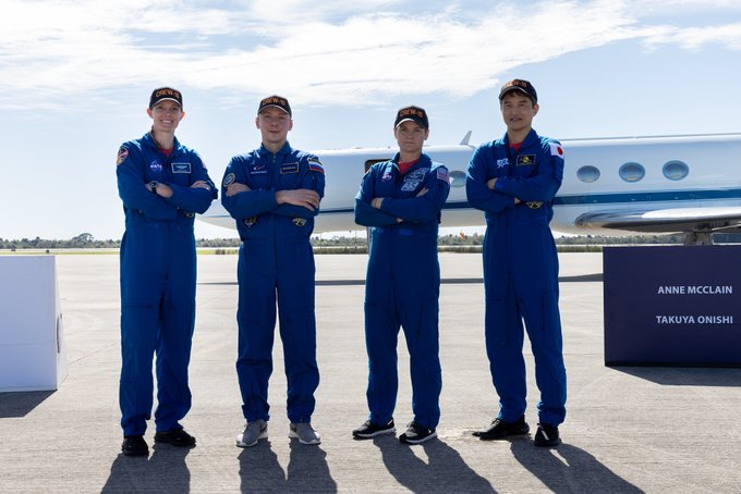 The four members of NASA's SpaceX Crew-10 mission — Nichole Ayers, Kirill Peskov, Anne McClain, and Takuya Onishi — stand on the tarmac at NASA's Kennedy Space Center following their arrival for launch. The four explorers are wearing blue flight suits and facing the camera with their arms crossed. In the background is the plane that brought them to Kennedy and a sign with Anne and Takuya's names visible. Credit: NASA/Kim Shiflett