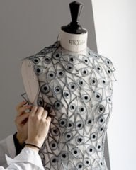 A designer meticulously works on an intricate, lace-like dress adorned with delicate patterns, displayed on a mannequin in a bright studio.