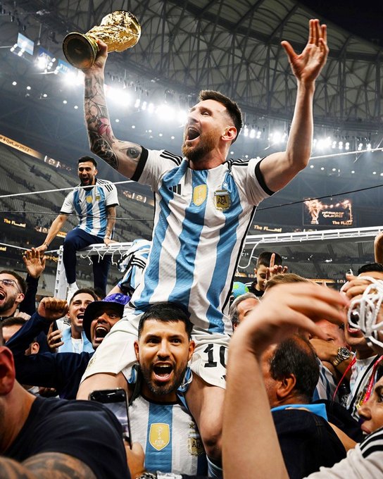 The image captures a jubilant moment of celebration, featuring a man, likely Lionel Messi, being carried on the shoulders of teammates while holding a golden trophy aloft. The setting is a large stadium filled with cheering fans, indicating a significant victory. The man is wearing an Argentina national football team jersey, signifying the team's qualification for the 2026 World Cup. The post text from Diario Olé highlights Messi's potential to be the first player to participate in six World Cups, noting he will be 39 years old during the tournament. The atmosphere is electric, with fans and players alike expressing immense joy and pride.
