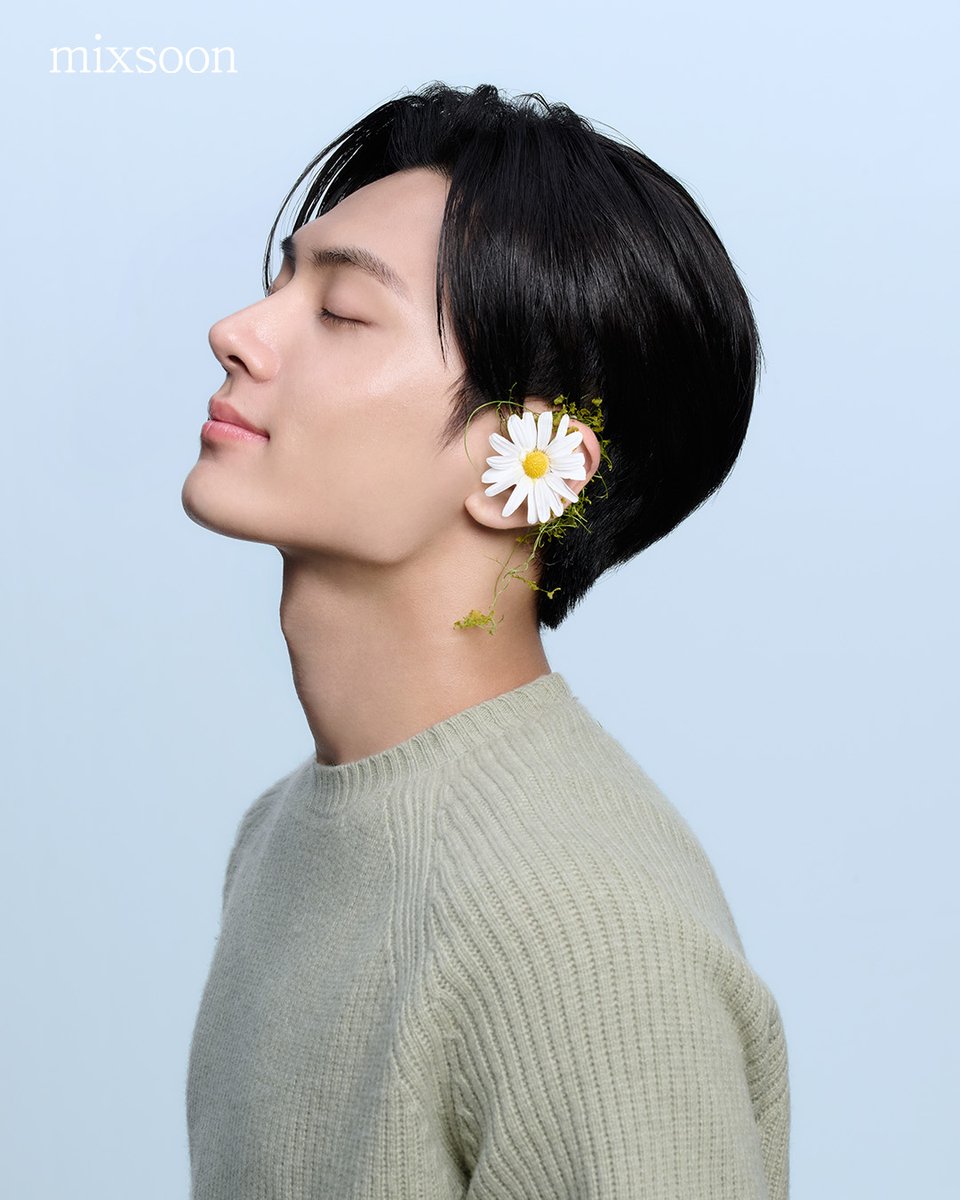 He sits there, a daisy tucked into his hair, and that soft green sweater against the calming blue backdrop, the first photoshoot for NEW BRAND AMBASSADOR, Mixsoon. There's this serene aura about him, like he's effortlessly living that Mixsoon 'radiant, natural beauty' vibe.