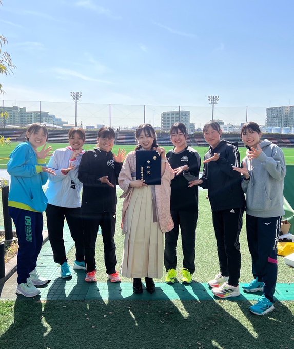 The first image captures a graduation ceremony at Tokyo Budokan, as indicated by the post text. A young woman, presumably Azusa, the team captain mentioned in the post, is standing in front of a large vertical banner with Japanese text that reads "帝京科学大学 卒業式" (Teikyo University of Science Graduation Ceremony). She is dressed in traditional academic attire, holding a diploma, and smiling warmly, reflecting the celebratory mood of the event. The setting is outdoors, with modern architectural elements visible in the background. The second image shows a group of young women, likely members of the Teikyo University of Science Women's Ekiden Team, posing together on a sports field. They are dressed in athletic wear, with one individual in the center also in graduation attire, holding a diploma. The group's cheerful expressions and hand gestures suggest camaraderie and support, celebrating the graduation of their teammate. The post text conveys gratitude towards Azusa for her leadership and wishes her success in her future endeavors in a corporate team.