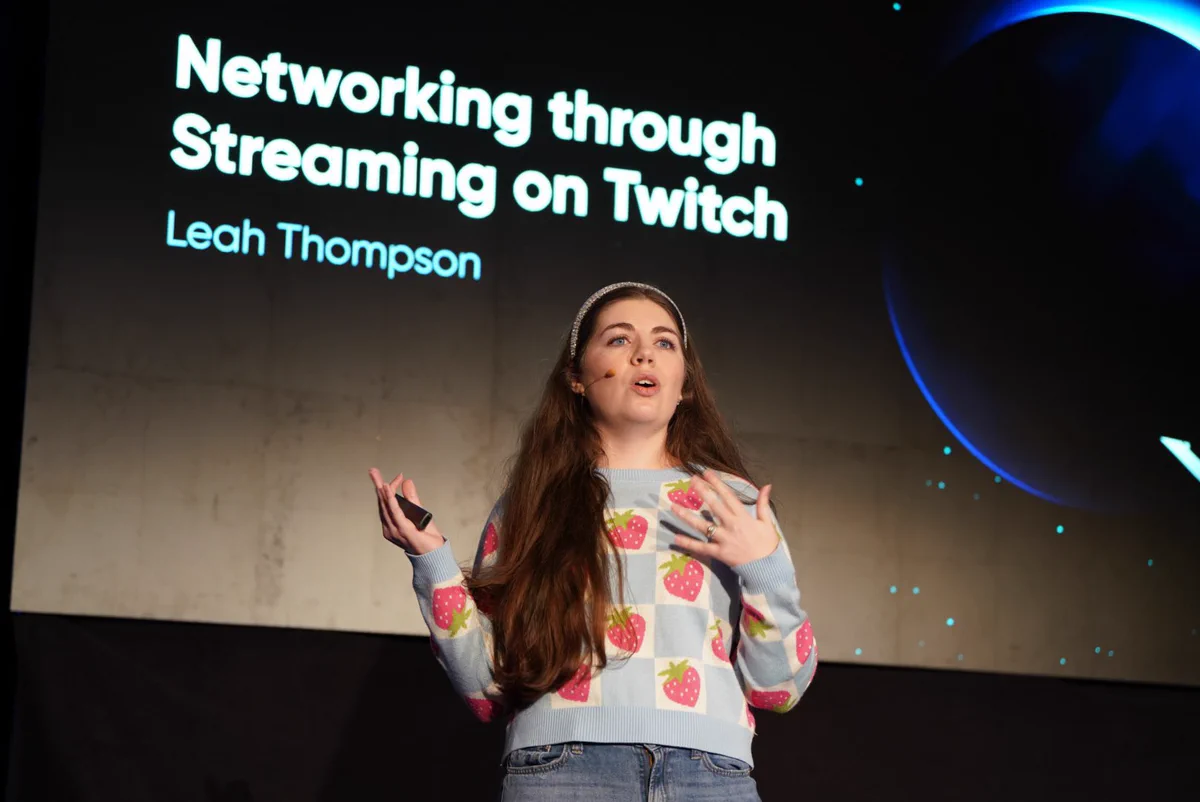 Leah, a woman with long brown hair and blue eyes, is standing on a stage. Behind her is a screen that says “Networking through streaming on Twitch”.