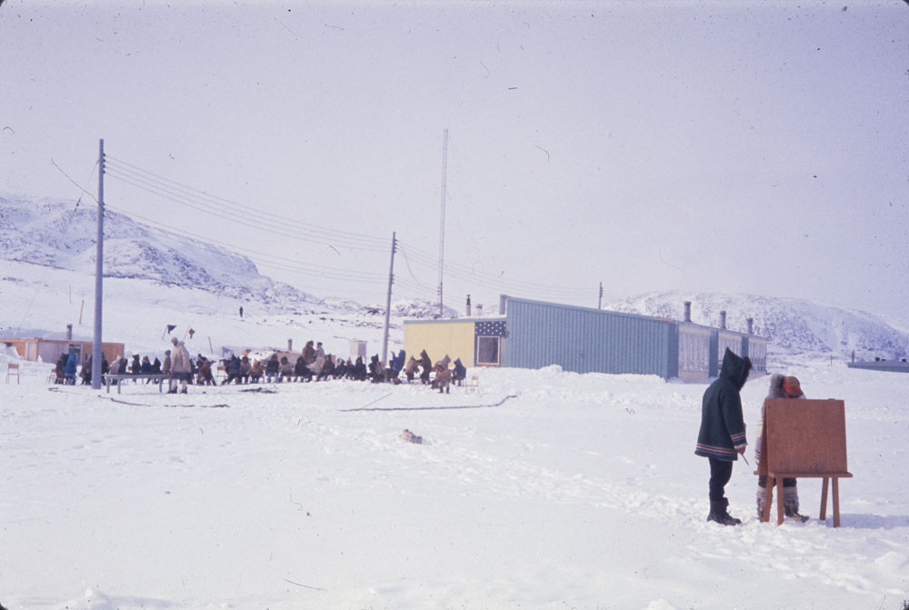 Two men check shooting scores as children play musical chairs in Kinngait, Nunavut.

Photo by Charles Gimpel, 1964. Likely April 24.