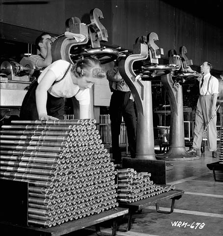 Woman stacks Bren gun barrels while workmen perform quality control. John Inglis Bren Plant in Toronto, Ontario. 

National Film Board. Taken April 8, 1941.