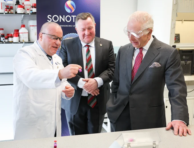 A scientist in a lab coat shows a small vial to two men in a lab. King Charles III wears a suit and tie, and the other wears a suit with safety goggles. They are in a laboratory setting with shelves of bottles and equipment in the background.