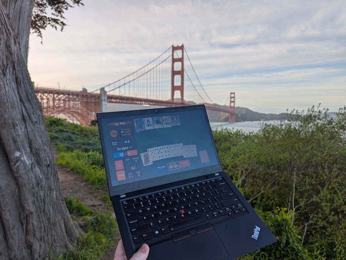Lenovo ThinkPad T495s running balatro in the foreground, being held up by my left hand. The Golden Gate Bridge is in the background 