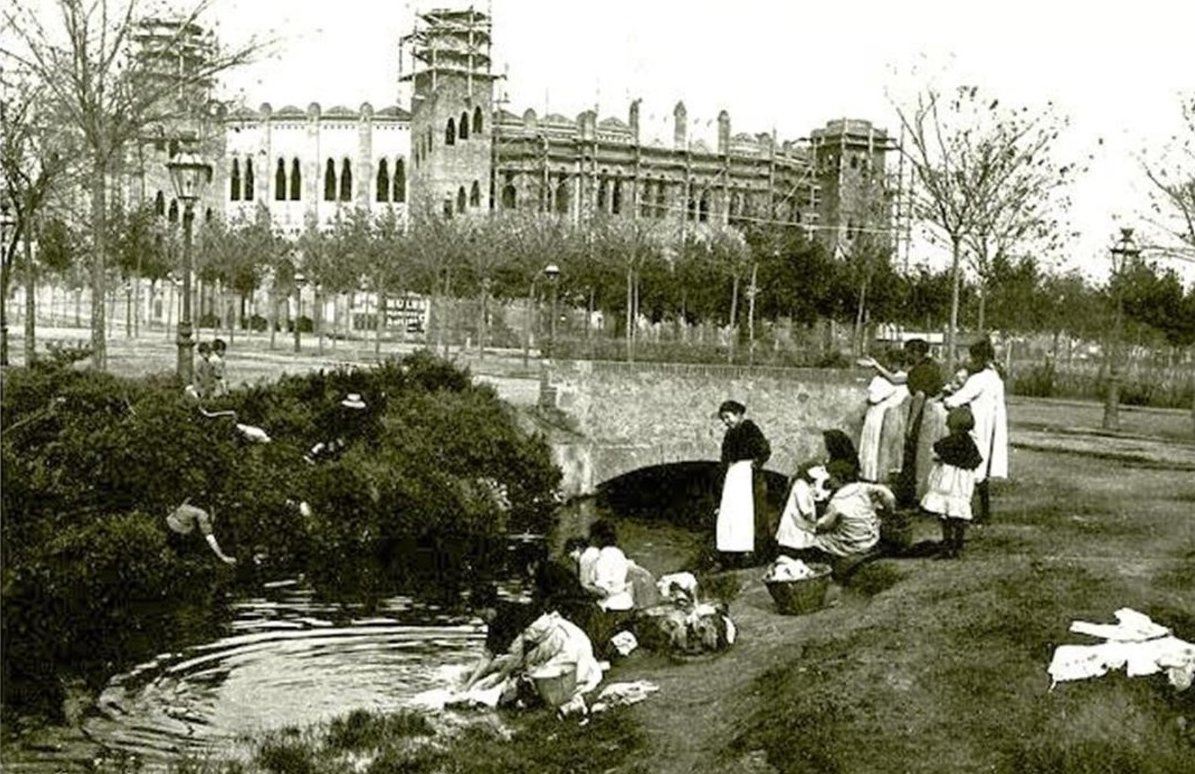 Un grup de dones fent a la bugada al Rec Comtal, al  carrer Marina amb Gran Via (c. 1915). 

Al fons, la Monumental en construcció.