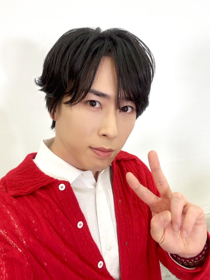 The image features Ryota Miyadate, a member of the Japanese idol group Snow Man, wearing a red cardigan over a white shirt. He is making a peace sign with his right hand, which is a common gesture in Japanese pop culture to convey friendliness and positivity. The post announces the release of Snow Man's album 'THE BEST 2020 - 2025' on streaming platforms, containing 61 songs. The text encourages fans to listen to the music and make it a part of their daily lives, asking which songs are their favorites. The overall tone of the post is celebratory and engaging, aimed at fans of the group.