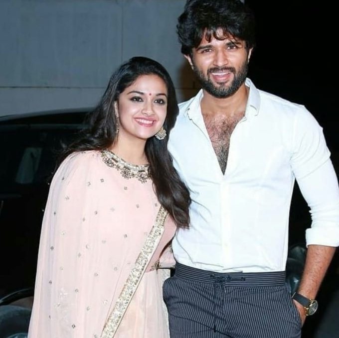 The image features two individuals, identified as Vijay Devarakonda and Keerthy Suresh, standing side by side. Vijay Devarakonda is dressed in a white shirt with rolled-up sleeves and dark trousers, while Keerthy Suresh is wearing a traditional light pink saree adorned with intricate embroidery. Both are smiling warmly, suggesting a friendly and positive atmosphere. The post text from Milagro Movies indicates that this is an exclusive image related to the project "RowdyJanardhan," hinting at a promotional or celebratory context. The background is neutral, focusing attention on the individuals, and the overall composition conveys a sense of camaraderie and professional collaboration.