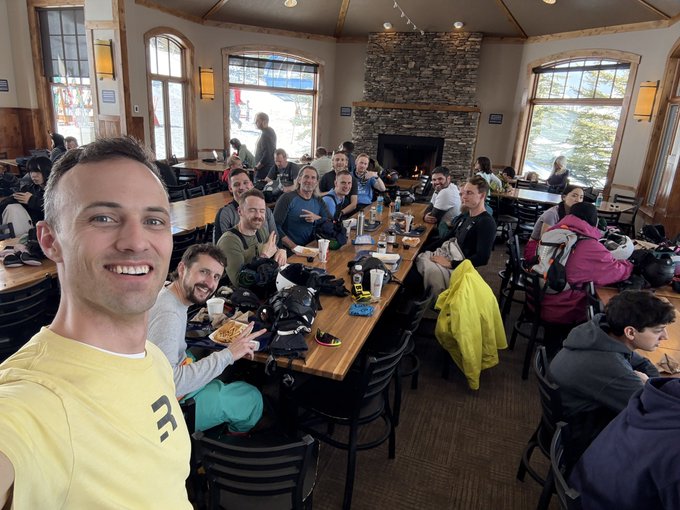 A group of around fifteen people sits around two long wooden tables inside a cozy ski lodge with large windows, a tall stone fireplace, and vaulted ceilings. In the foreground, a man in a yellow shirt takes a cheerful selfie with others smiling, chatting, and eating food like fries and burgers. Ski helmets, gloves, and gear are scattered across the tables. Outside the windows, snow-covered slopes and ski lifts are visible, adding to the lively après-ski atmosphere.