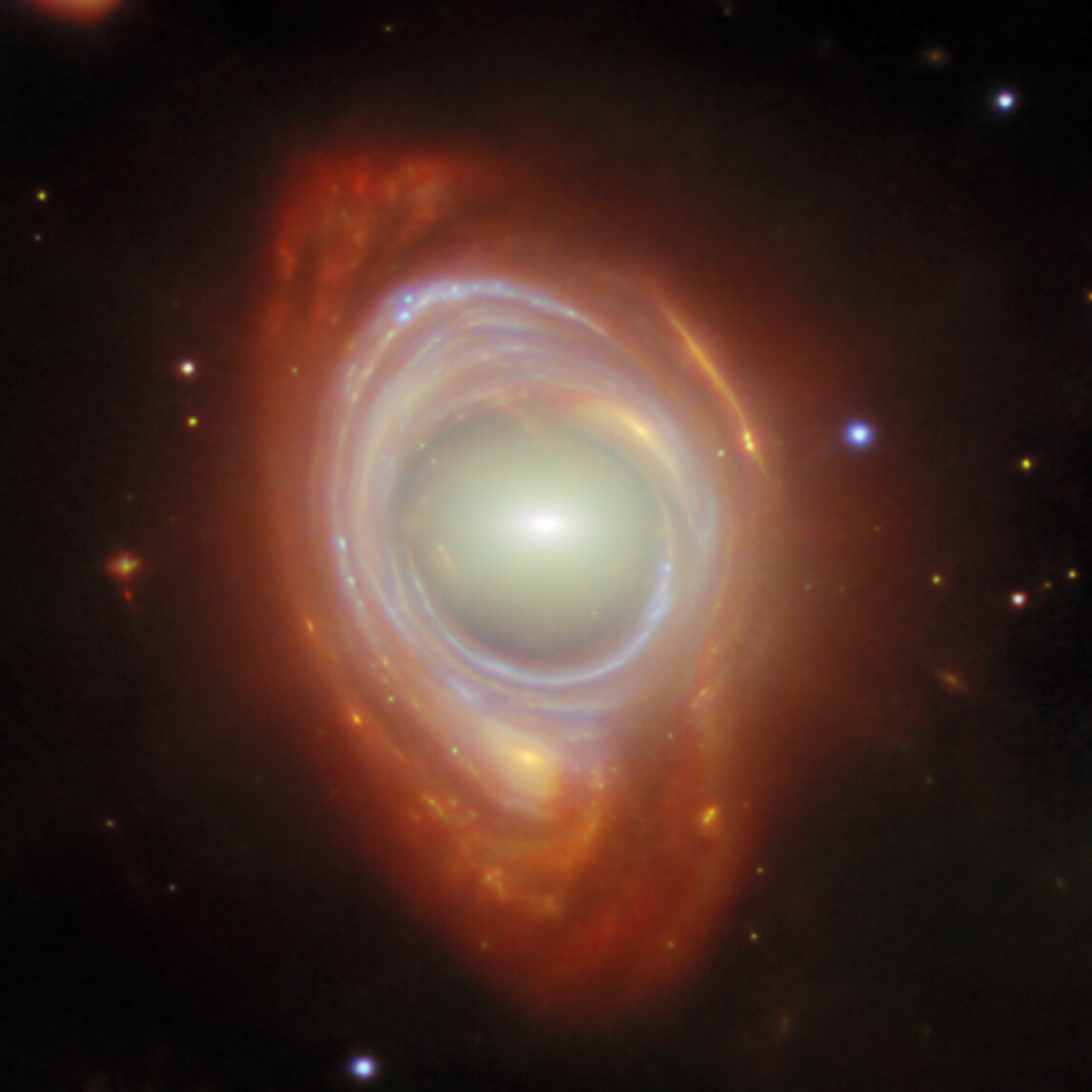In the center is an elliptical galaxy, seen as an oval-shaped glow around a small bright core. Around this is wrapped a broad band of light, appearing like a spiral galaxy stretched and warped into a ring, with bright blue lines drawn through it where the spiral arms have been stretched into circles. A few distant objects are visible around the ring on a black background. Credit: ESA/Webb, NASA & CSA, G. Mahler