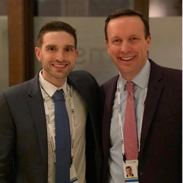 The image shows two men in business attire, wearing lanyards with name badges, suggesting they are at a professional or political conference. The man on the right is identified as a Connecticut senator, likely Chris Murphy, based on the context provided in the original post text. The setting appears to be indoors, possibly at a formal event or meeting. The post text references a conspiracy theory involving the senator working with organizations to overthrow the president, mentioning Serbian Otpor, which is associated with CIA and USAID-led regime change efforts. This context adds a layer of political intrigue and controversy to the image.
