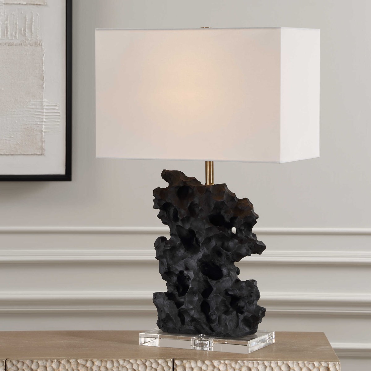 Crafted from a black man-made stone, this unique table lamp replicates a rock sculpture that boasts heavy texture with distinctive holes, displayed atop a thick crystal base with plated antique brass hardware. #carolynkinder #uttermost uttermost.com/basalt-table-l…