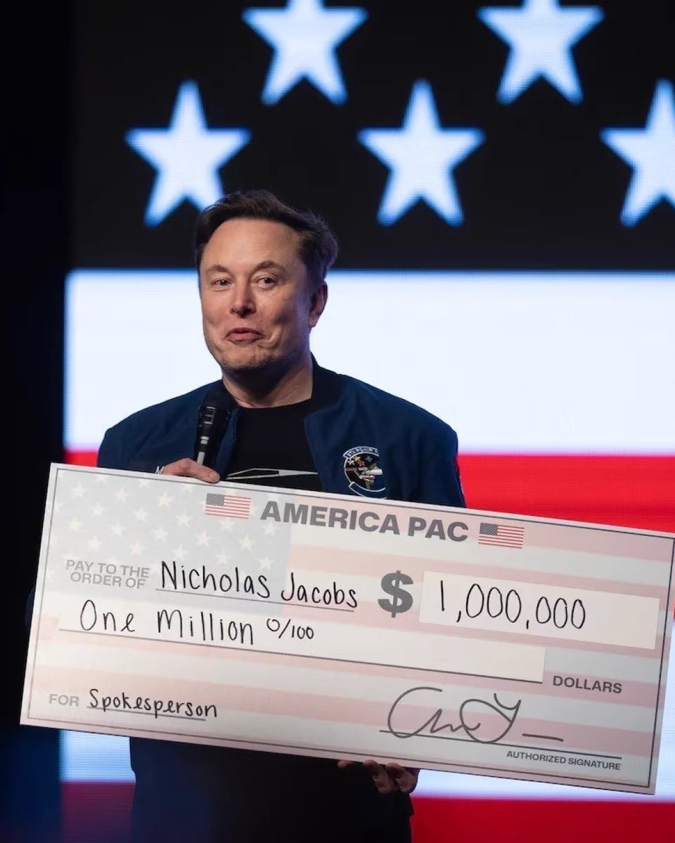 Elon Musk stands on stage holding a large ceremonial check from America PAC addressed to Nicholas Jacobs for one million dollars, with a backdrop featuring stars.