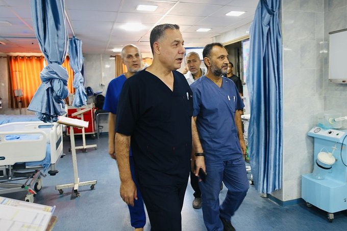 Palestinian human rights defender Dr. Hussam Abu Safiya, wearing dark blue medical scrubs, walks through a hospital ward with other medical staff. Hospital beds, blue privacy curtains, and medical equipment are visible in the background.