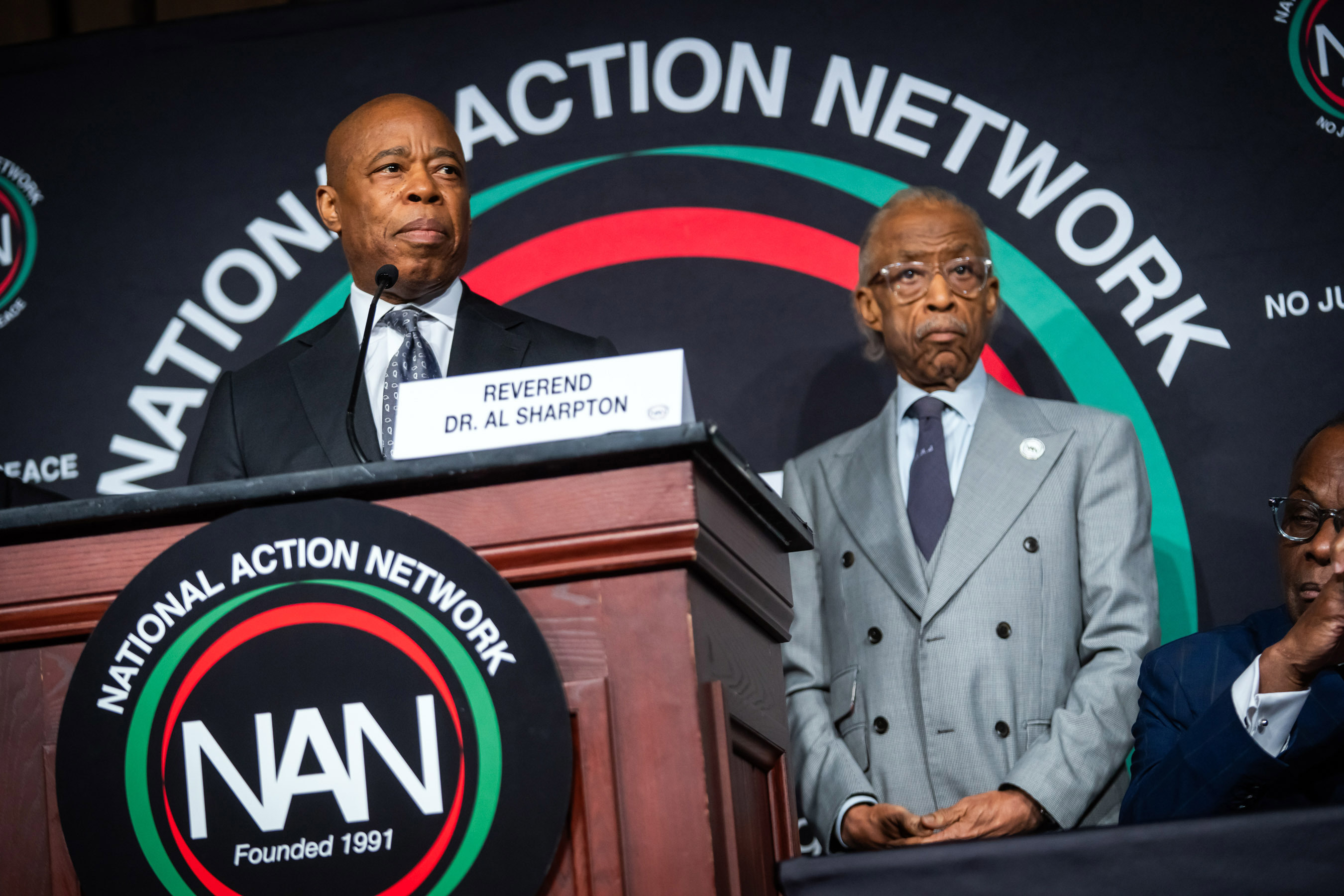 mayor stands at podium to give remarks with rev al sharpton behind him and sign behind them both that reads national action network