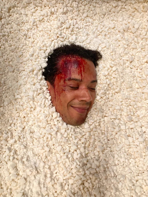 Jacob Anderson’s head, his forehead covered in SFX blood, peaks out from a bed of rocks with eyes closed and a slight smile. 