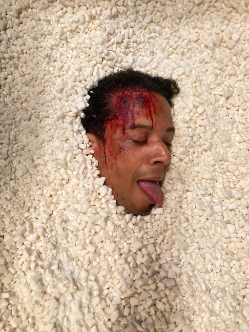 Jacob Anderson’s head, his forehead covered in SFX blood, peaks out from a bed of rocks with eyes closed and his tongue stuck out. 