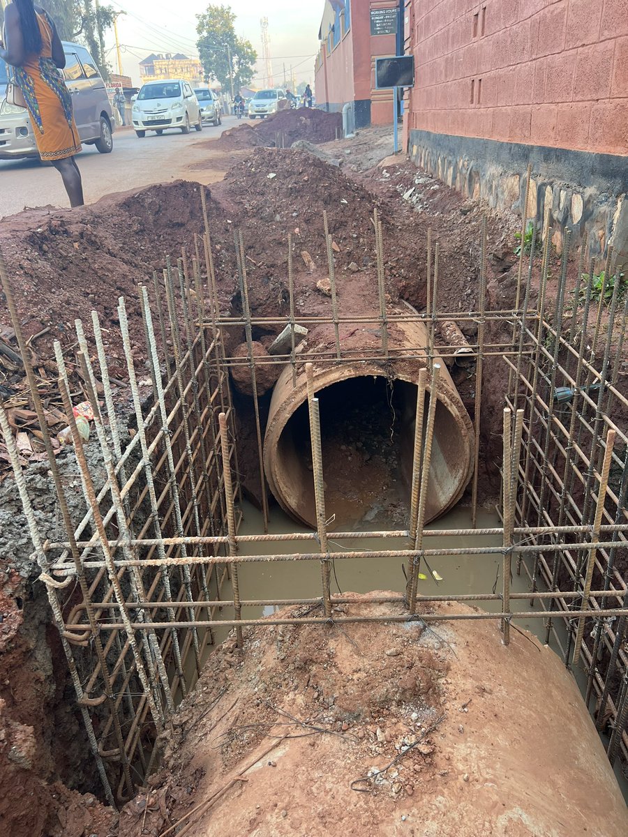 ApolloBuregyeya's tweet photo. KAMPALA: Where Gravity is Optional and Sewage is Freelance
==========
Dear @KCCAED, 
I know you’re Muslim, but there are three ways of drinking Uganda Waragi: with water, without water, and like water.

Unfortunately, it seems our urban engineers in Kyebando chose the third option before designing the drainage system.

What else explains the confident burial of a massive concrete pipe in a trench with zero gradient, as if stormwater will flow through sheer optimism? The pipe lies there like a retired civil servant, wide, experienced, but completely inert.

Here, let’s talk use case. You see, this pipe isn’t just for stormwater — it also moonlights as a neighborhood sewage transporter. When it rains, latrines and septic tanks perched proudly above send their greetings downstream. That’s what we call functional urban innovation. You know it’s real? But instead of acknowledging this unholy alliance of rain and refuse, we’re installing a closed pipe system that pretends the people are flushing poetry and rose petals.

Then the other failure: the disappearing act of maintenance. The old open drain, for all its unattractiveness, allowed for quick interventions. If it blocked, someone saw it, and someone fixed it (usually barefoot). Now? We’ve sealed the mess in a basement with no windows, no access, and no maintenance plan. Out of sight, out of mind, until it overflows into someone’s house during Sunday lunch.

Next? Social context blindness. Behavior isn’t an Excel formula. If your design doesn’t match how people live (if you ignore grey water, waste habits, and solid disposal patterns), you’re building fantasy, not infrastructure.

Lastly, we’ve declared war on physics. Water doesn’t care about political will or PR photos. If it can’t flow through your pipe, it’ll flow on your road. And when sewage and stormwater start using tarmac as a runway, potholes will bloom, kids will get sick, and the very road you upgraded becomes a microbial slipway.

So. Before we close another drainage system in Kampala, can we:

1. Model the flow — with actual data, not just gut feelings?

2. Study what’s entering the drain — because it’s not just rain, it’s real life?

3. Design with access in mind — so when it clogs (not if), someone can clear it?

4. Accept behavioral realities — and build for how people actually use infrastructure? 

5. Respect the laws of gravity — because the Lord gave them for free?

Or we can keep drinking Waragi like water… and building drains that do the same. The engineers on this project need to be found and given a medal of dishonor, @KCCAUG @UIPE_Uganda @UACE_Uganda