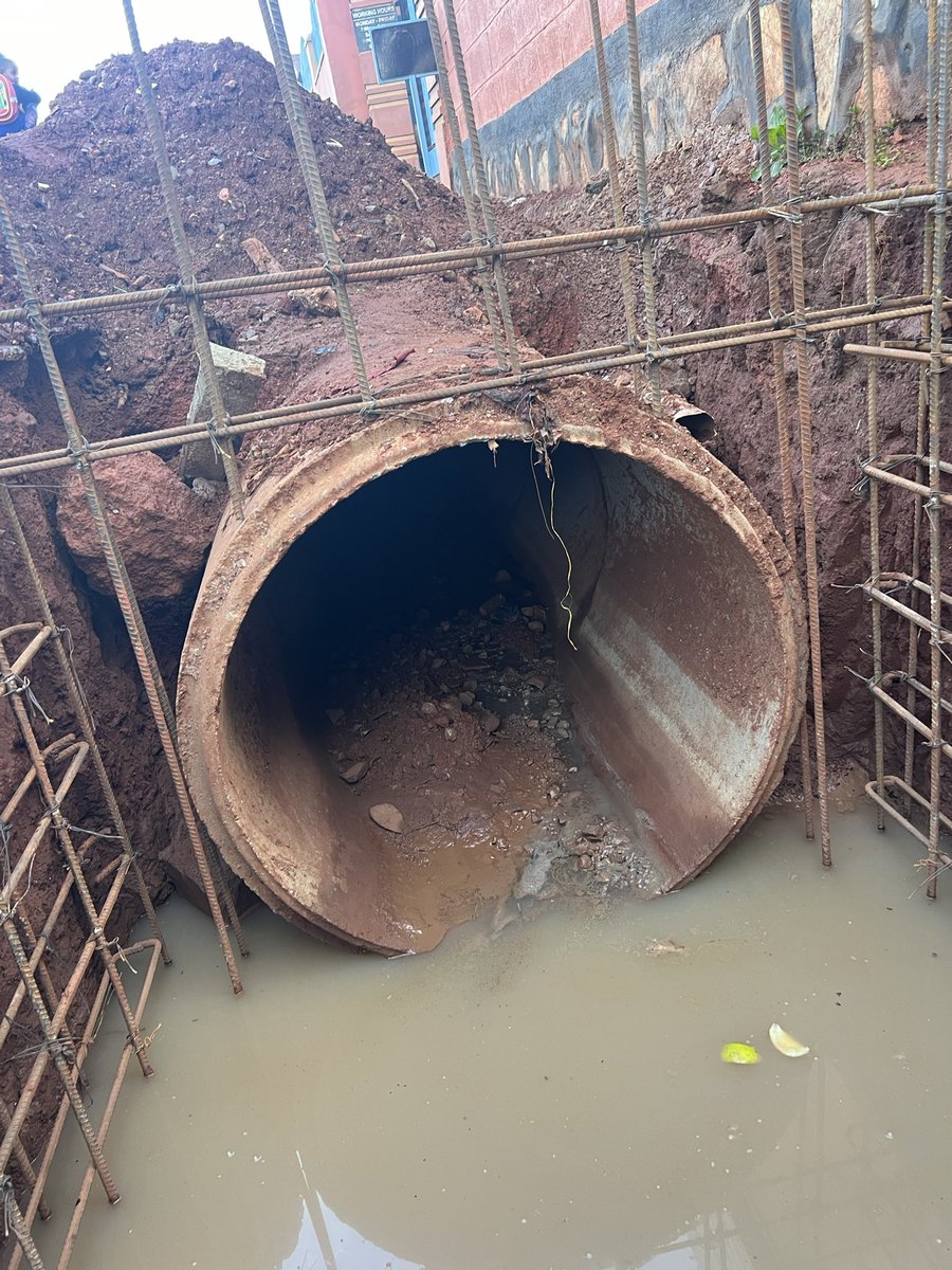 ApolloBuregyeya's tweet photo. KAMPALA: Where Gravity is Optional and Sewage is Freelance
==========
Dear @KCCAED, 
I know you’re Muslim, but there are three ways of drinking Uganda Waragi: with water, without water, and like water.

Unfortunately, it seems our urban engineers in Kyebando chose the third option before designing the drainage system.

What else explains the confident burial of a massive concrete pipe in a trench with zero gradient, as if stormwater will flow through sheer optimism? The pipe lies there like a retired civil servant, wide, experienced, but completely inert.

Here, let’s talk use case. You see, this pipe isn’t just for stormwater — it also moonlights as a neighborhood sewage transporter. When it rains, latrines and septic tanks perched proudly above send their greetings downstream. That’s what we call functional urban innovation. You know it’s real? But instead of acknowledging this unholy alliance of rain and refuse, we’re installing a closed pipe system that pretends the people are flushing poetry and rose petals.

Then the other failure: the disappearing act of maintenance. The old open drain, for all its unattractiveness, allowed for quick interventions. If it blocked, someone saw it, and someone fixed it (usually barefoot). Now? We’ve sealed the mess in a basement with no windows, no access, and no maintenance plan. Out of sight, out of mind, until it overflows into someone’s house during Sunday lunch.

Next? Social context blindness. Behavior isn’t an Excel formula. If your design doesn’t match how people live (if you ignore grey water, waste habits, and solid disposal patterns), you’re building fantasy, not infrastructure.

Lastly, we’ve declared war on physics. Water doesn’t care about political will or PR photos. If it can’t flow through your pipe, it’ll flow on your road. And when sewage and stormwater start using tarmac as a runway, potholes will bloom, kids will get sick, and the very road you upgraded becomes a microbial slipway.

So. Before we close another drainage system in Kampala, can we:

1. Model the flow — with actual data, not just gut feelings?

2. Study what’s entering the drain — because it’s not just rain, it’s real life?

3. Design with access in mind — so when it clogs (not if), someone can clear it?

4. Accept behavioral realities — and build for how people actually use infrastructure? 

5. Respect the laws of gravity — because the Lord gave them for free?

Or we can keep drinking Waragi like water… and building drains that do the same. The engineers on this project need to be found and given a medal of dishonor, @KCCAUG @UIPE_Uganda @UACE_Uganda