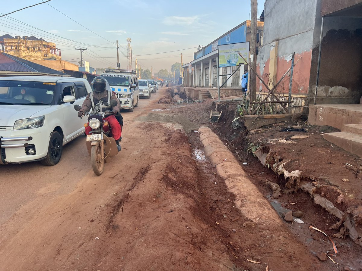 ApolloBuregyeya's tweet photo. KAMPALA: Where Gravity is Optional and Sewage is Freelance
==========
Dear @KCCAED, 
I know you’re Muslim, but there are three ways of drinking Uganda Waragi: with water, without water, and like water.

Unfortunately, it seems our urban engineers in Kyebando chose the third option before designing the drainage system.

What else explains the confident burial of a massive concrete pipe in a trench with zero gradient, as if stormwater will flow through sheer optimism? The pipe lies there like a retired civil servant, wide, experienced, but completely inert.

Here, let’s talk use case. You see, this pipe isn’t just for stormwater — it also moonlights as a neighborhood sewage transporter. When it rains, latrines and septic tanks perched proudly above send their greetings downstream. That’s what we call functional urban innovation. You know it’s real? But instead of acknowledging this unholy alliance of rain and refuse, we’re installing a closed pipe system that pretends the people are flushing poetry and rose petals.

Then the other failure: the disappearing act of maintenance. The old open drain, for all its unattractiveness, allowed for quick interventions. If it blocked, someone saw it, and someone fixed it (usually barefoot). Now? We’ve sealed the mess in a basement with no windows, no access, and no maintenance plan. Out of sight, out of mind, until it overflows into someone’s house during Sunday lunch.

Next? Social context blindness. Behavior isn’t an Excel formula. If your design doesn’t match how people live (if you ignore grey water, waste habits, and solid disposal patterns), you’re building fantasy, not infrastructure.

Lastly, we’ve declared war on physics. Water doesn’t care about political will or PR photos. If it can’t flow through your pipe, it’ll flow on your road. And when sewage and stormwater start using tarmac as a runway, potholes will bloom, kids will get sick, and the very road you upgraded becomes a microbial slipway.

So. Before we close another drainage system in Kampala, can we:

1. Model the flow — with actual data, not just gut feelings?

2. Study what’s entering the drain — because it’s not just rain, it’s real life?

3. Design with access in mind — so when it clogs (not if), someone can clear it?

4. Accept behavioral realities — and build for how people actually use infrastructure? 

5. Respect the laws of gravity — because the Lord gave them for free?

Or we can keep drinking Waragi like water… and building drains that do the same. The engineers on this project need to be found and given a medal of dishonor, @KCCAUG @UIPE_Uganda @UACE_Uganda
