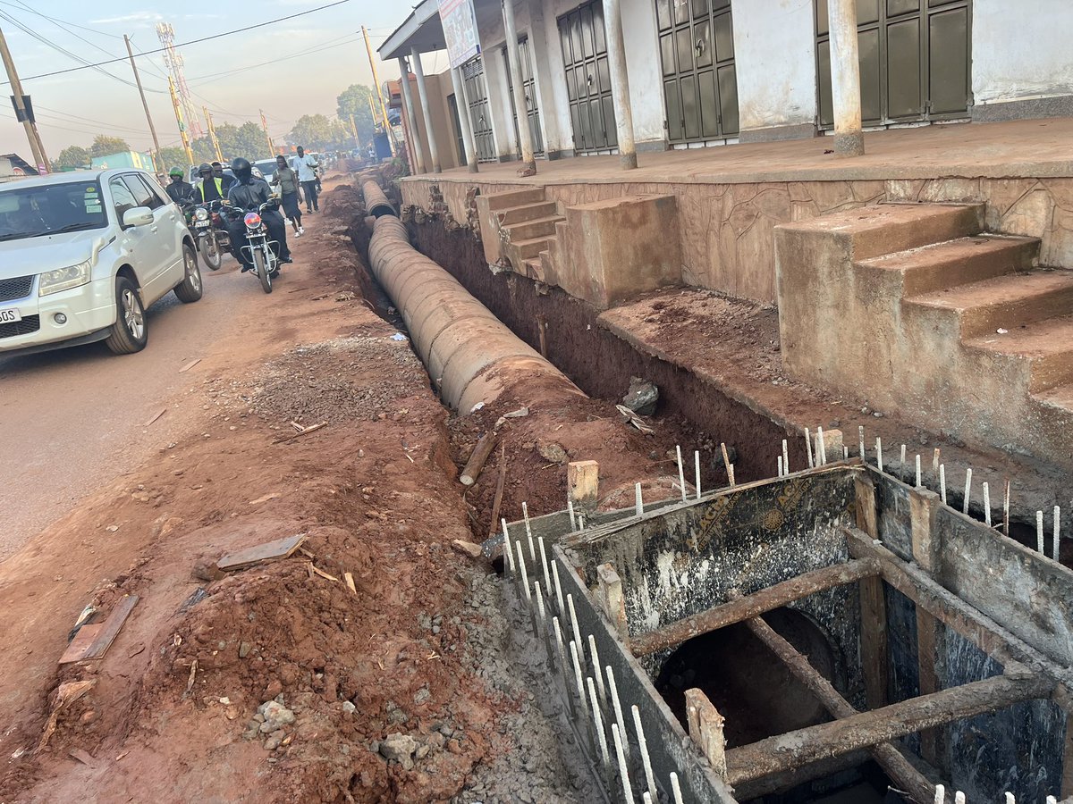 ApolloBuregyeya's tweet photo. KAMPALA: Where Gravity is Optional and Sewage is Freelance
==========
Dear @KCCAED, 
I know you’re Muslim, but there are three ways of drinking Uganda Waragi: with water, without water, and like water.

Unfortunately, it seems our urban engineers in Kyebando chose the third option before designing the drainage system.

What else explains the confident burial of a massive concrete pipe in a trench with zero gradient, as if stormwater will flow through sheer optimism? The pipe lies there like a retired civil servant, wide, experienced, but completely inert.

Here, let’s talk use case. You see, this pipe isn’t just for stormwater — it also moonlights as a neighborhood sewage transporter. When it rains, latrines and septic tanks perched proudly above send their greetings downstream. That’s what we call functional urban innovation. You know it’s real? But instead of acknowledging this unholy alliance of rain and refuse, we’re installing a closed pipe system that pretends the people are flushing poetry and rose petals.

Then the other failure: the disappearing act of maintenance. The old open drain, for all its unattractiveness, allowed for quick interventions. If it blocked, someone saw it, and someone fixed it (usually barefoot). Now? We’ve sealed the mess in a basement with no windows, no access, and no maintenance plan. Out of sight, out of mind, until it overflows into someone’s house during Sunday lunch.

Next? Social context blindness. Behavior isn’t an Excel formula. If your design doesn’t match how people live (if you ignore grey water, waste habits, and solid disposal patterns), you’re building fantasy, not infrastructure.

Lastly, we’ve declared war on physics. Water doesn’t care about political will or PR photos. If it can’t flow through your pipe, it’ll flow on your road. And when sewage and stormwater start using tarmac as a runway, potholes will bloom, kids will get sick, and the very road you upgraded becomes a microbial slipway.

So. Before we close another drainage system in Kampala, can we:

1. Model the flow — with actual data, not just gut feelings?

2. Study what’s entering the drain — because it’s not just rain, it’s real life?

3. Design with access in mind — so when it clogs (not if), someone can clear it?

4. Accept behavioral realities — and build for how people actually use infrastructure? 

5. Respect the laws of gravity — because the Lord gave them for free?

Or we can keep drinking Waragi like water… and building drains that do the same. The engineers on this project need to be found and given a medal of dishonor, @KCCAUG @UIPE_Uganda @UACE_Uganda