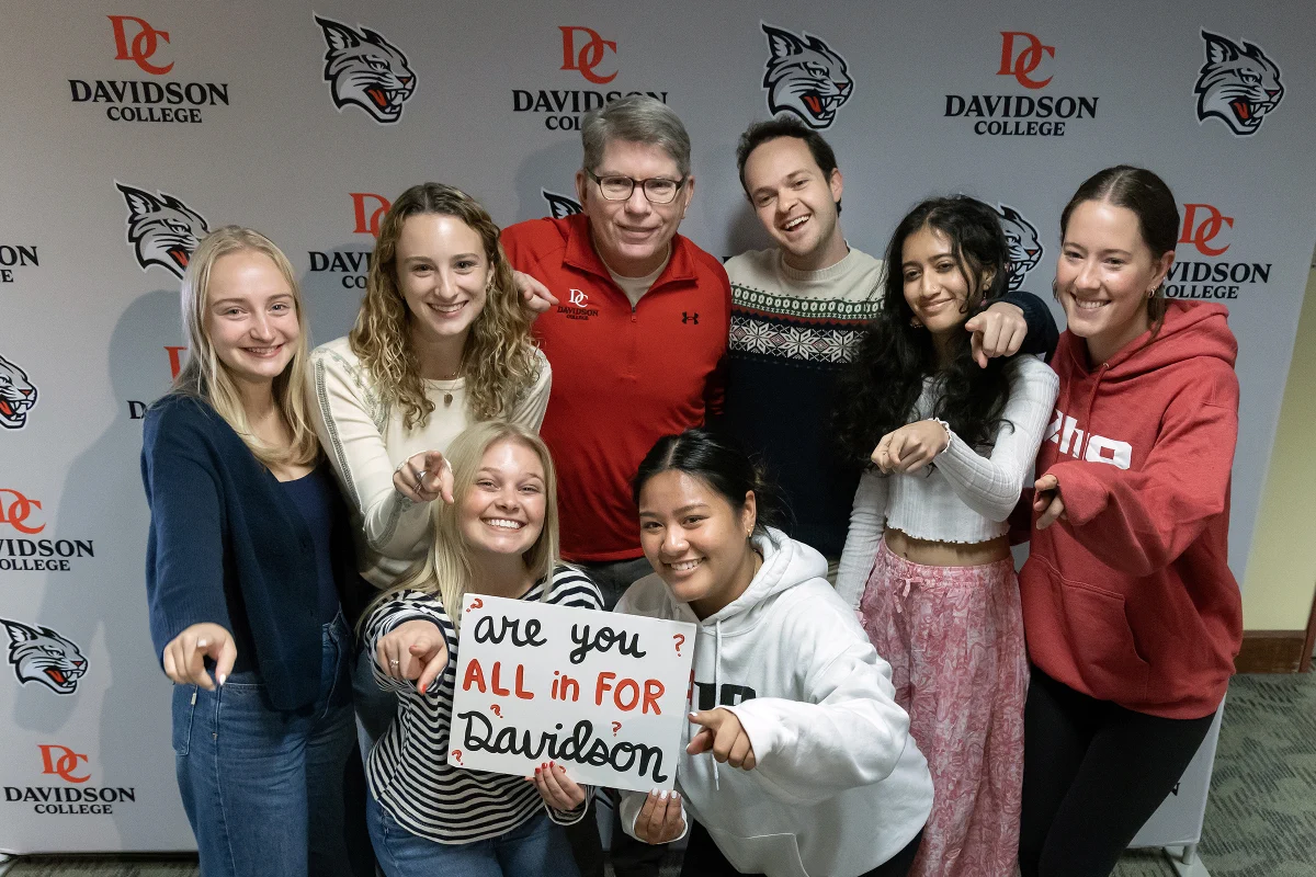A photo of President Doug Hicks standing and smiling with a group of students in front of a Davidson College backdrop.