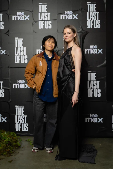 Picture at The Last of Us photo wall with Gabby and Anne. Gabby is wearing a tan The Last of Us Carhartt jacket. Anne is wearing a black satin dress with barbed wire detailing.
