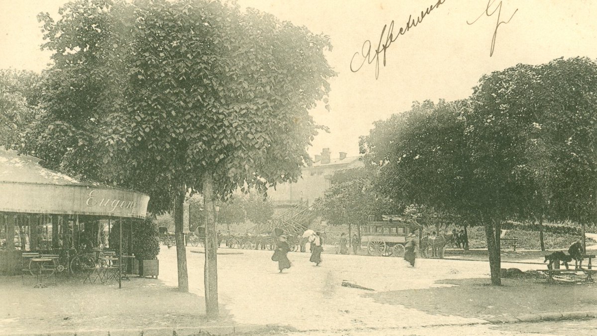 Place de l’arrivée (place Gasnier Guy), gare de Chelles. 
Au fond, le bâtiment de la gare, l’escalier descendant des quais, une calèche qui attend les voyageurs. À gauche une brasserie ; le lieu un temps occupé par une librairie-papeterie a retrouvé sa vocation d’origine.